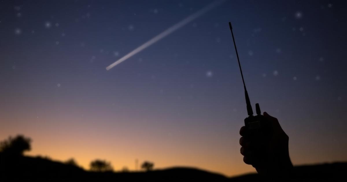 Silhouette of a hand holding an HT skyward at night with a satellite streak overhead